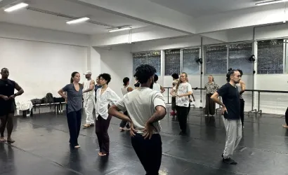 Grupo de bailarinos em uma ampla sala de ensaio com piso preto linóleo e paredes brancas iluminadas por luzes de led. Os dançarinos vestem roupas confortáveis e casuais em tons neutros — como branco, preto e cinza — e estão espalhados pelo espaço, alguns de costas e outros de frente, praticando movimentos corporais. Ao fundo, observam-se grandes janelas e barras de dança encostadas na parede.