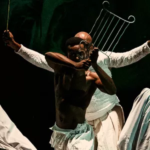 Imagem enquadra um rapaz negro, à frente, que segura um indumentária do orixá Iroko chamado "Orelha do Tempo" (ou grelha). Atrás dele, um homem negro mais velho está enquadrado apenas com a metade da cabeça, com o olhar por trás do rapaz e ele segura, nas duas mãos, folhas da espada de Ogum.