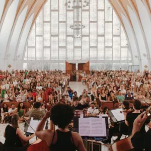 Imagem enquadra músicos da OSBA posicionados em um palco para frente de um público de pessoas sentadas em bancos dentro de um Santuário 