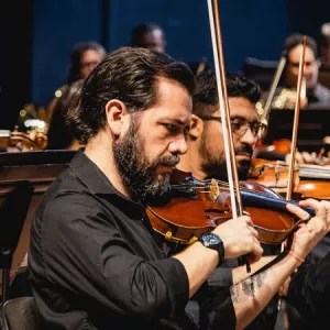 Imagem centraliza, ao meio, um músico com um violino. Ele é um homem branco, de barba, e um relógio no braço. Ele está de perfil. Em volta dele estão outros músicos, homens, que também estão com violinos. Todos usam camisa social preta.