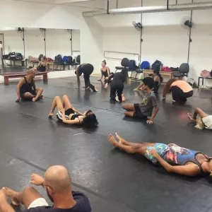 Imagem mostra um grupo de pessoas reunidas em uma sala de ensaio de dança. Algumas estão deitadas, outras estão em posição de alongamento.