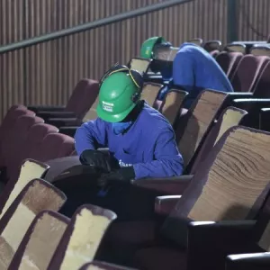 Imagem enquadra dois trabalhadores que utilizam capacetes verdes. Eles estão retirando poltronas da Sala Principal do Teatro Castro Alves