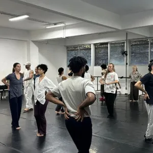 Grupo de bailarinos em uma ampla sala de ensaio com piso preto linóleo e paredes brancas iluminadas por luzes de led. Os dançarinos vestem roupas confortáveis e casuais em tons neutros — como branco, preto e cinza — e estão espalhados pelo espaço, alguns de costas e outros de frente, praticando movimentos corporais. Ao fundo, observam-se grandes janelas e barras de dança encostadas na parede.