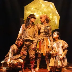 Quatro atores em uma peça de teatro, vestidos com roupas retalhadas em tons de terra, no palco escuro. No centro, dois deles, com narizes postiços, estão sob um guarda-chuva amarelo iluminado por pequenas luzes. Uma terceira atriz, agachada à esquerda, e uma atriz em pé à direita olham atentamente para cima.