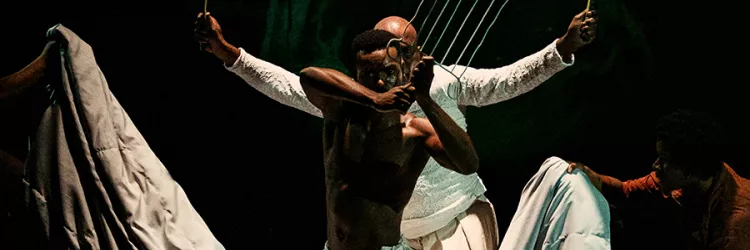 Imagem enquadra um rapaz negro, à frente, que segura um indumentária do orixá Iroko chamado "Orelha do Tempo" (ou grelha). Atrás dele, um homem negro mais velho está enquadrado apenas com a metade da cabeça, com o olhar por trás do rapaz e ele segura, nas duas mãos, folhas da espada de Ogum.