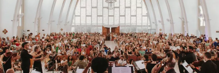 Imagem enquadra músicos da OSBA posicionados em um palco para frente de um público de pessoas sentadas em bancos dentro de um Santuário 