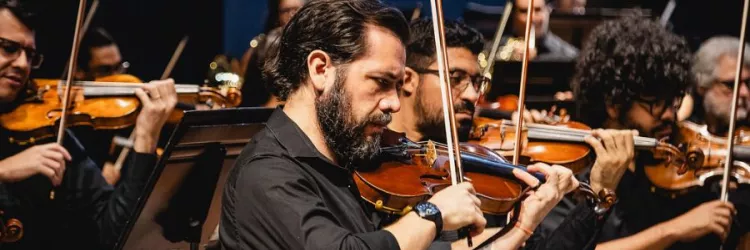 Imagem centraliza, ao meio, um músico com um violino. Ele é um homem branco, de barba, e um relógio no braço. Ele está de perfil. Em volta dele estão outros músicos, homens, que também estão com violinos. Todos usam camisa social preta.