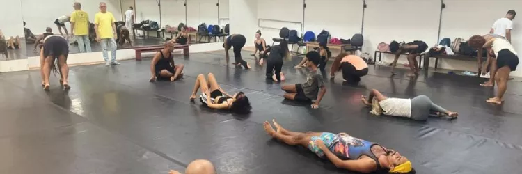 Imagem mostra um grupo de pessoas reunidas em uma sala de ensaio de dança. Algumas estão deitadas, outras estão em posição de alongamento.