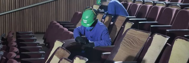 Imagem enquadra dois trabalhadores que utilizam capacetes verdes. Eles estão retirando poltronas da Sala Principal do Teatro Castro Alves