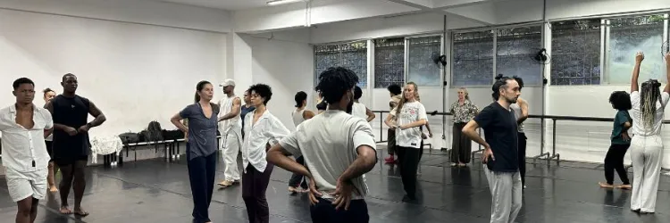 Grupo de bailarinos em uma ampla sala de ensaio com piso preto linóleo e paredes brancas iluminadas por luzes de led. Os dançarinos vestem roupas confortáveis e casuais em tons neutros — como branco, preto e cinza — e estão espalhados pelo espaço, alguns de costas e outros de frente, praticando movimentos corporais. Ao fundo, observam-se grandes janelas e barras de dança encostadas na parede.
