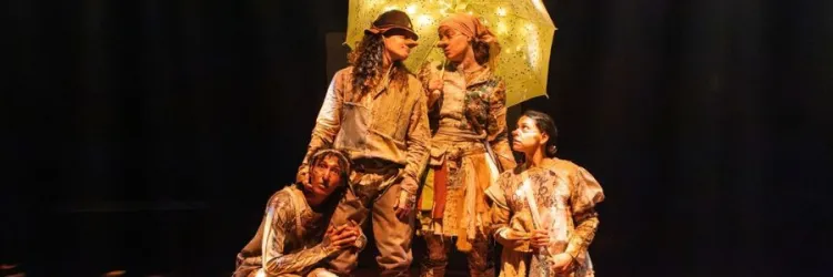 Quatro atores em uma peça de teatro, vestidos com roupas retalhadas em tons de terra, no palco escuro. No centro, dois deles, com narizes postiços, estão sob um guarda-chuva amarelo iluminado por pequenas luzes. Uma terceira atriz, agachada à esquerda, e uma atriz em pé à direita olham atentamente para cima.