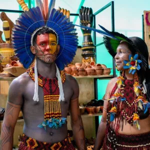 duas pessoas indígenas, um homem e uma mulher, em frente a artesanatos